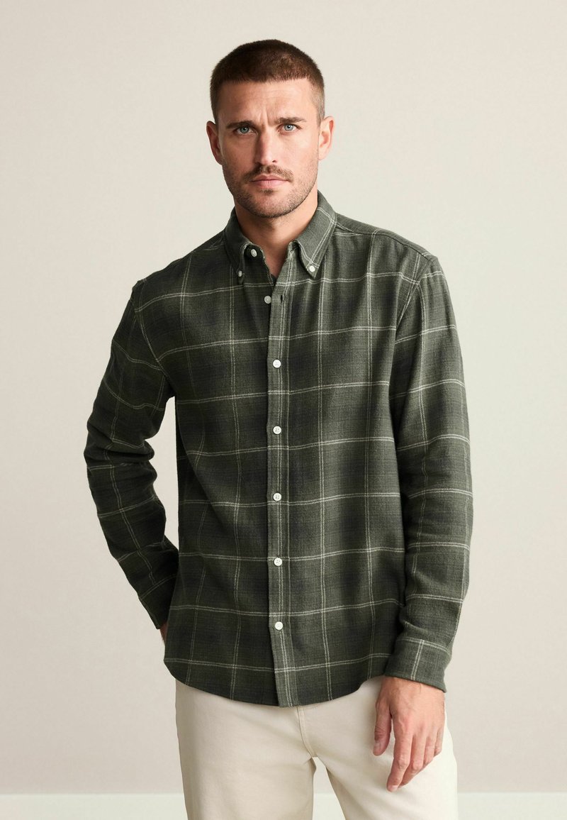 Camisa de franela de cuadros en verde oscuro con un patrón de cuadrícula blanca, cuello de botón y botones blancos. Mangas largas con puños ajustados y bajo recto.