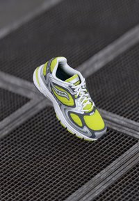 Gray and white running shoe with yellow mesh upper, intricate design, reflective accents, and cushioned sole, positioned on a textured surface.