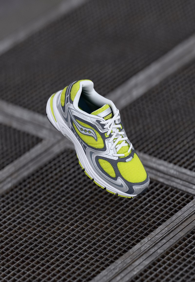 Gray and white running shoe with yellow mesh upper, intricate design, reflective accents, and cushioned sole, positioned on a textured surface.