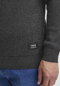 Maglione grigio scuro realizzato in tessuto morbido con polsini e orlo a costine. Presenta un'etichetta nera con testo bianco nell'angolo in basso a sinistra.