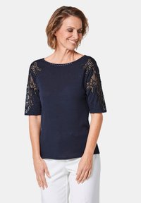 Donkerblauw topje met korte mouwen, voorzien van doorschijnende geborduurde kant met florale ontwerpen en subtiele versieringen langs de halslijn.