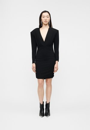 Nő áll egy egyszínű fehér háttér előtt, fekete, vállpántos, hosszú ujjú ruhát viselve, mély V-nyakkal és fekete bokacsizmával.