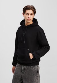 Čierna mikina s kapucňou, predným vreckom, šnúrkou na stiahnutie a malou vzorovanou nášivkou. Vyrobená z mäkkej látky a s rebrovanými manžetami a spodným lemom.
