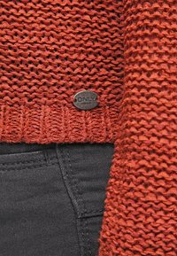 Pull en tricot couleur rouille avec une étiquette métallique de la marque "ONLY" au-dessus de la poche d'un jean denim noir, montrant la texture du tricot et les détails de la couture.