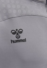 Czarne logo hummel na jasnoszarej tkaninie sportowej z fakturowanym wzorem kropek w pobliżu szwu.