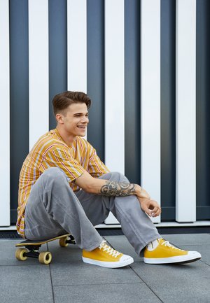 Gele sneakers met witte zolen, grijze wijde broekspijpen en een gestreept geel-wit shirt. Persoon zit op een skateboard tegen een muur.