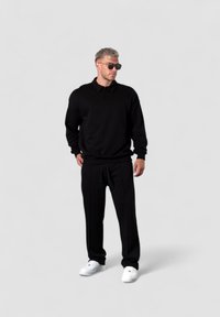 Schwarzer Pullover mit einem Reißverschlusskragen kombiniert mit schwarzen Jogginghosen. Weiße Sneakers runden das Outfit ab. Glatte Textur insgesamt.