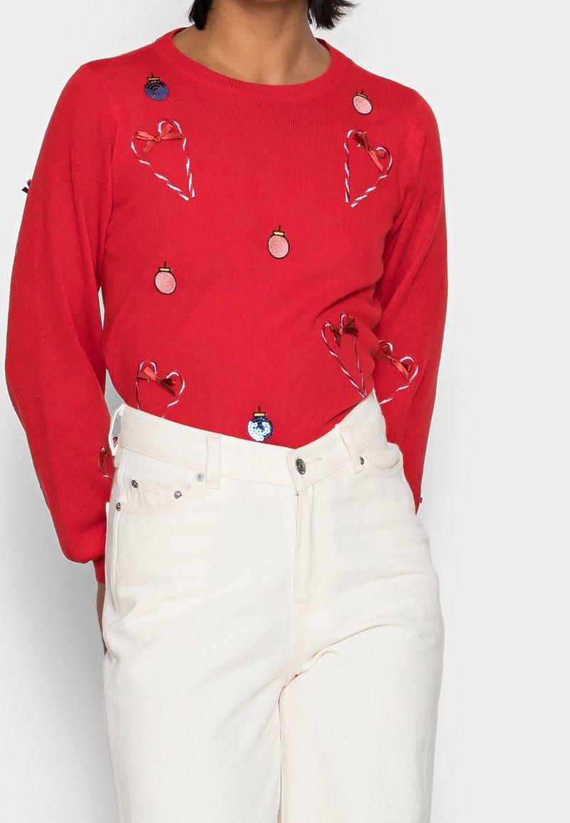 Rode gebreide trui met geborduurde ornamenten, zuurstokken en harten. Gecombineerd met witte jeans met hoge taille en zilveren knopen.