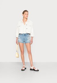 Jaqueta de ganga branca sobre uma blusa clara com bolinhas, calções de ganga rasgados e sandálias bicolores brancas e pretas. Bolsa amarela na mão.