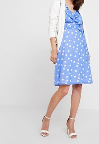 Robe bleue à pois avec un décolleté volanté et un design à enrouler. Portée avec un cardigan blanc et des talons blancs à lanières. Texture de tissu lisse.