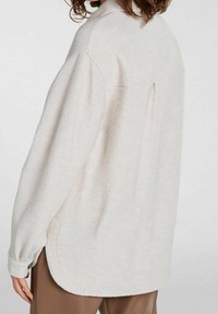 Kobieta nosząca luźną, jasnobeżową, teksturowaną koszulę z długim rękawem, z zaokrąglonym dołem i przeszyciami z tyłu, w połączeniu z brązowymi spodniami.