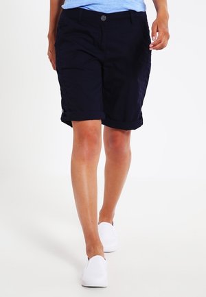 Person iført marineblå shorts med op-rullede kanter, lyseblå top og hvide slip-on sko, der går fremad på en hvid baggrund.