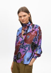 Blusa floreale con sfondo viola, caratterizzata da grandi fiori rossi, blu e neri. Tessuto leggero, maniche lunghe e dettaglio con fiocco al collo.