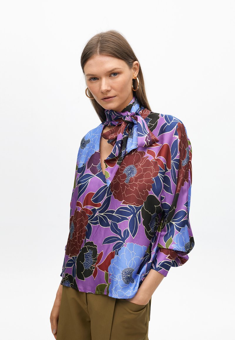 Blusa floreale con sfondo viola, caratterizzata da grandi fiori rossi, blu e neri. Tessuto leggero, maniche lunghe e dettaglio con fiocco al collo.