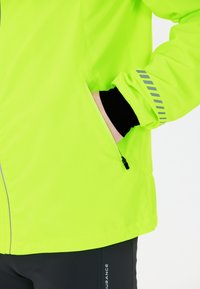 Neonvihreä takki, jossa on vetoketjullinen tasku, joustavat hihansuut ja heijastavat harmaat raidat, yhdistettynä mustiin alaosiin. Kevyt ja sileä rakenne.