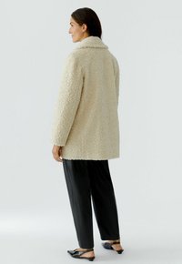 Cream, textured wool coat with a collar and long sleeves, paired with black loose-fitting pants and flat shoes.