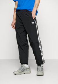 Svarta träningsbyxor med vita sidostråk, elastisk midja och fickor med dragkedja. Bärs med grå sportskor. Slät tygtextur.