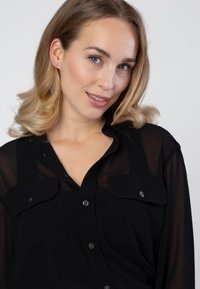 Zwarte doorschijnende button-up blouse met twee voorzakken, voorzien van een klassieke kraag en subtiele knoopaccenten. Model heeft licht golvend haar.