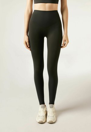 Donna che indossa leggings neri a vita alta e scarpe da ginnastica bianche, in piedi con le braccia rilassate ai lati su uno sfondo semplice.