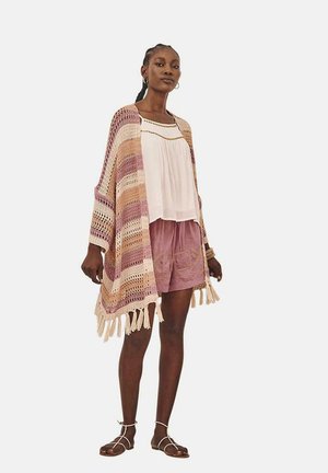 Poncho de ganchillo en rosa y beige con un dobladillo con flecos, superpuesto a una blusa blanca y pantalones cortos rosas, con patrones texturizados.