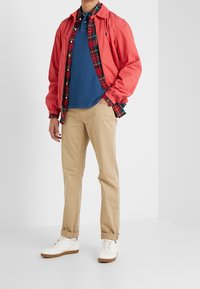 Man med korallfärgad jacka, röd rutiga skjorta, blå polo, beige byxor och vita sneakers står i ett vitt rum med händerna i fickorna.