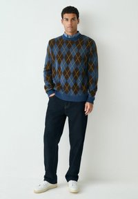 Gestreifter Argyle-Pullover in Blau, Braun und Gelb; Rundhalsausschnitt, gerippte Bündchen. Kombiniert mit dunklen Jeans und weißen Sneakers.