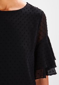 Blusa nera in tessuto testurizzato con un delicato motivo a pois. Ha maniche corte arricciate e un collo rotondo.