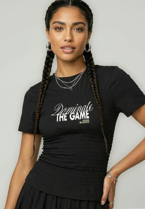 Žena s vrkočmi, ktorá nosí strieborné náušnice kruhy, vrstvené náhrdelníky, čierne tričko s nápisom „Dominate The Game“ a čiernu sukňu.