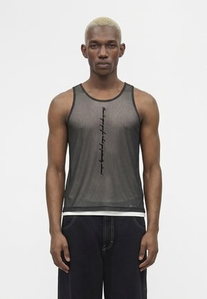 Muž s krátkymi blond vlasmi nosí čierne priehľadné tielko s vertikálnym čiernym textom a tmavé nohavice, stojí pred jednofarebným bielym pozadím.