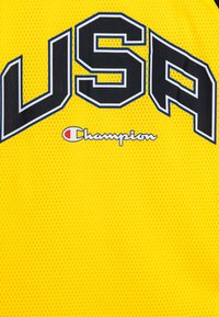 Maglia gialla in rete con grande scritta nera "USA" e contorni blu. Sotto, il logo "Champion" in bianco con un accento rosso.