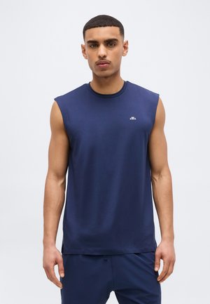 Férfi ujjatlan sötétkék sportfelsőben és hozzáillő nadrágban, semleges arckifejezéssel, egyszínű háttér előtt állva.