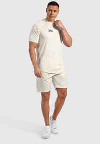 Camisa de manga corta beige con "SMILDOOD" impreso en negro, combinada con pantalones cortos a juego. Zapatillas blancas completan el atuendo atlético.