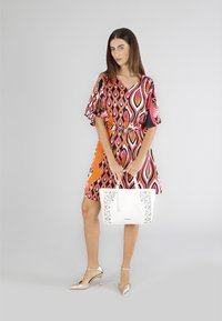 Abito multicolore con scollo a V e maniche a campana, accentuato da un pannello arancione. Borsa a tracolla bianca con design floreale ritagliato, tacchi dorati.