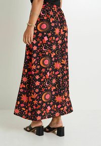 Falda maxi de tela negra con vibrantes patrones celestiales en rosa y naranja. Cintura elástica, diseño fluido, combinada con sandalias de tacón cuadrado negras.
