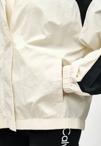 Χέρι στην τσέπη ενός μπουφάν με κρέμα και μαύρο φερμουάρ, σε συνδυασμό με μαύρα κολάν που δείχνουν μερικό λευκό κείμενο.