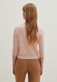 Jasnoróżowy, dziergany sweter z rękawami do połowy przedramienia i ściągaczem u do�łu, noszony na beżowych spodniach. Widok od tyłu ukazujący gładką fakturę i subtelne przeszycia.