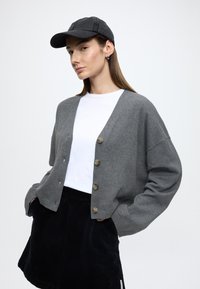 Šedý pletený kardigán s nadrozmernými rukávmi, päť drevených gombíkov, nosený cez biele tričko, v kombinácii s čiernou manšestrovou sukňou a šiltovkou.