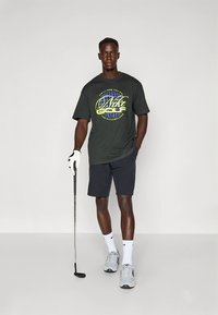 Juoda trumpomis rankovėmis marškinėliai su spalvingu „Nike Golf“ grafiniu dizainu, derinami su tamsiomis trumpomis kelnėmis, golfo pirštine ir pilkais sportbačiais ant modelio.