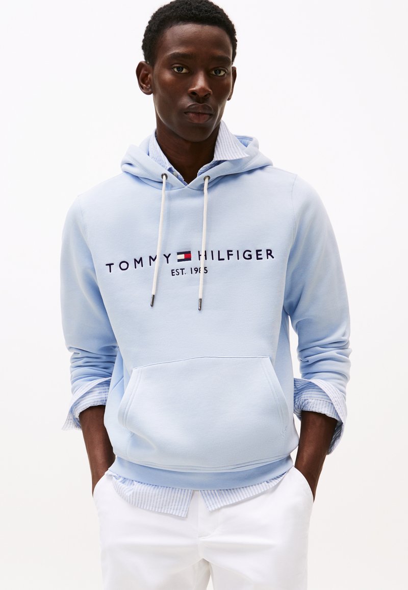 Hanorac din bumbac albastru deschis cu glugă, buzunar frontal, șnururi albe și logo "Tommy Hilfiger" cu litere albastre închise.