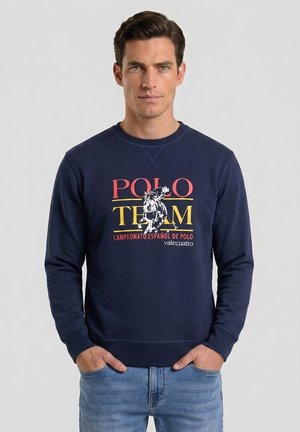 Hombre con sudadera azul marino con texto y gráfico "Polo Team Campeonato Español de Polo" en rojo y amarillo, manos en los bolsillos, vaqueros azules.