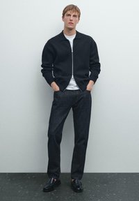 Junger Mann steht da, trägt eine schwarze Zip-Jacke, ein weißes T-Shirt, dunkle Jeans und schwarze Lederschuhe vor einer schlichten hellen Wand.