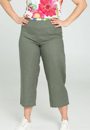 Personne portant un pantalon court vert olive, un haut floral coloré, des baskets blanches, debout les mains sur les hanches.