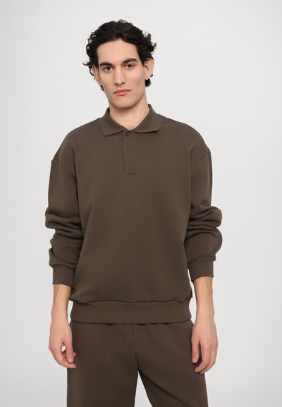Καφέ βαμβακερό φανελάκι τύπου πόλο με γιακά, λεπτομέρειες κουμπιών και τυλιγμένα μανίκια. Λείο υλικό και άνετη εφαρμογή. Συνδυάζεται με αντίστοιχες παντελόνες.