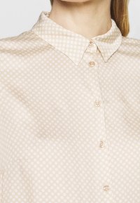 Beige button-up shirt with a subtle white polka dot pattern, featuring a classic collar and smooth fabric texture.
