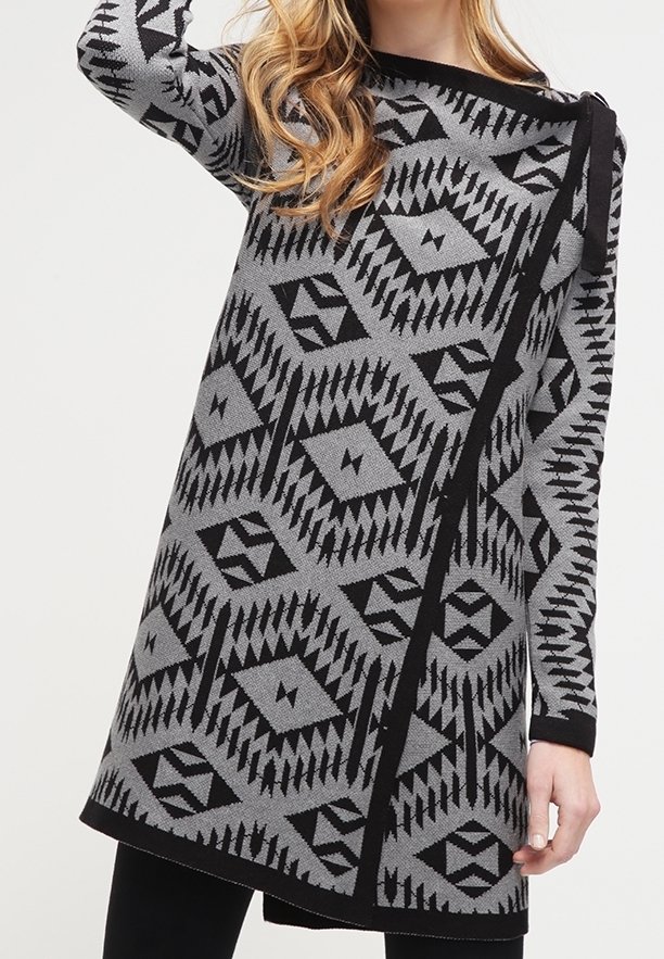 Robe tricotée grise avec des motifs géométriques noirs. Manches longues, ourlet asymétrique, et détail de nœud à l'épaule. Texture douce.