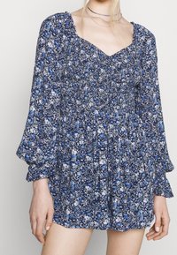 Modré květinové šaty se shromážděným korpusem a dlouhými, nadýchanými rukávy. Obsahují bílé a světle modré květiny na tmavě modrém pozadí.