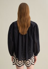 Dunkelbluse in Marineblau mit gepufften Ärmeln, strukturiertem Material und runder Kragen, kombiniert mit gemusterten Shorts in Cream und Schwarz.