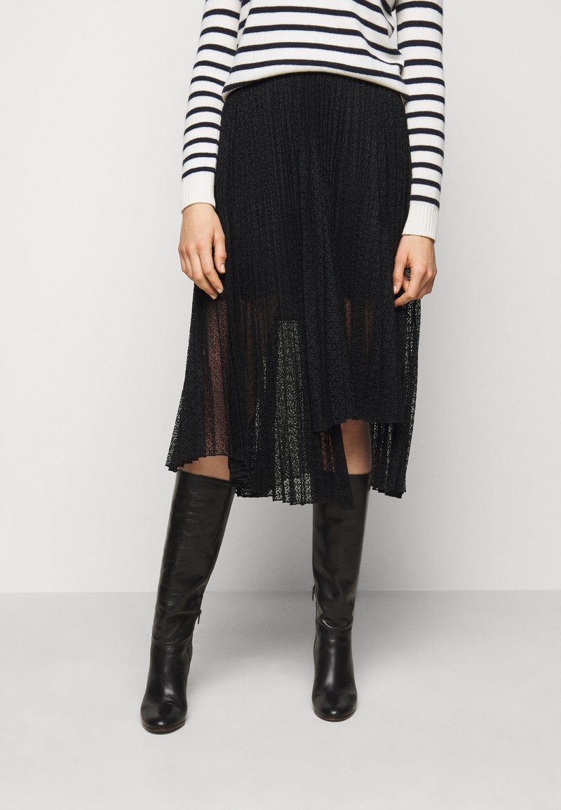 Femme portant une jupe midi plissée noire transparente, des bottes en cuir noires montantes jusqu'aux genoux, et un top à manches longues blanc et rayé bleu marine.