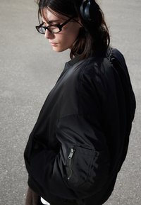Musta bomber-takki, jossa on vetoketjullinen tasku vasemmassa hihassa, sileä ja kevyt rakenne. Henkilö, jolla on lasit ja kuulokkeet.