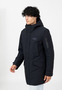 Schwarzer langer Mantel mit Kapuze, ausgestattet mit einem Reißverschluss vorne, Seitentaschen und einem Emblem auf der Brust. Glatte Textur mit einem lässigen Design.